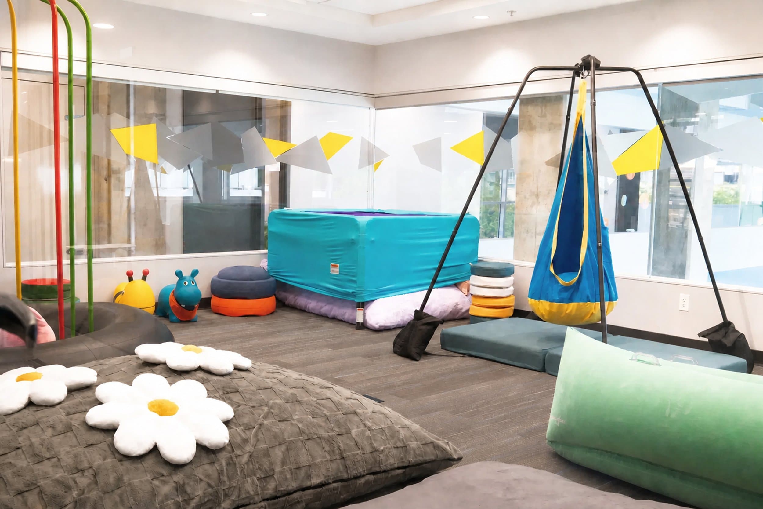 Sensory therapy room at The SPOT Pediatric Centrein Vancouver, BC, featuring a suspended platform swing, soft floor cushions, and floral and animal-shaped seating beneath large windows.